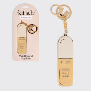 Gold metal enamel keychain depicting the kitsch Warm Sugar hair perfume with the packaging on the left on a light gray background