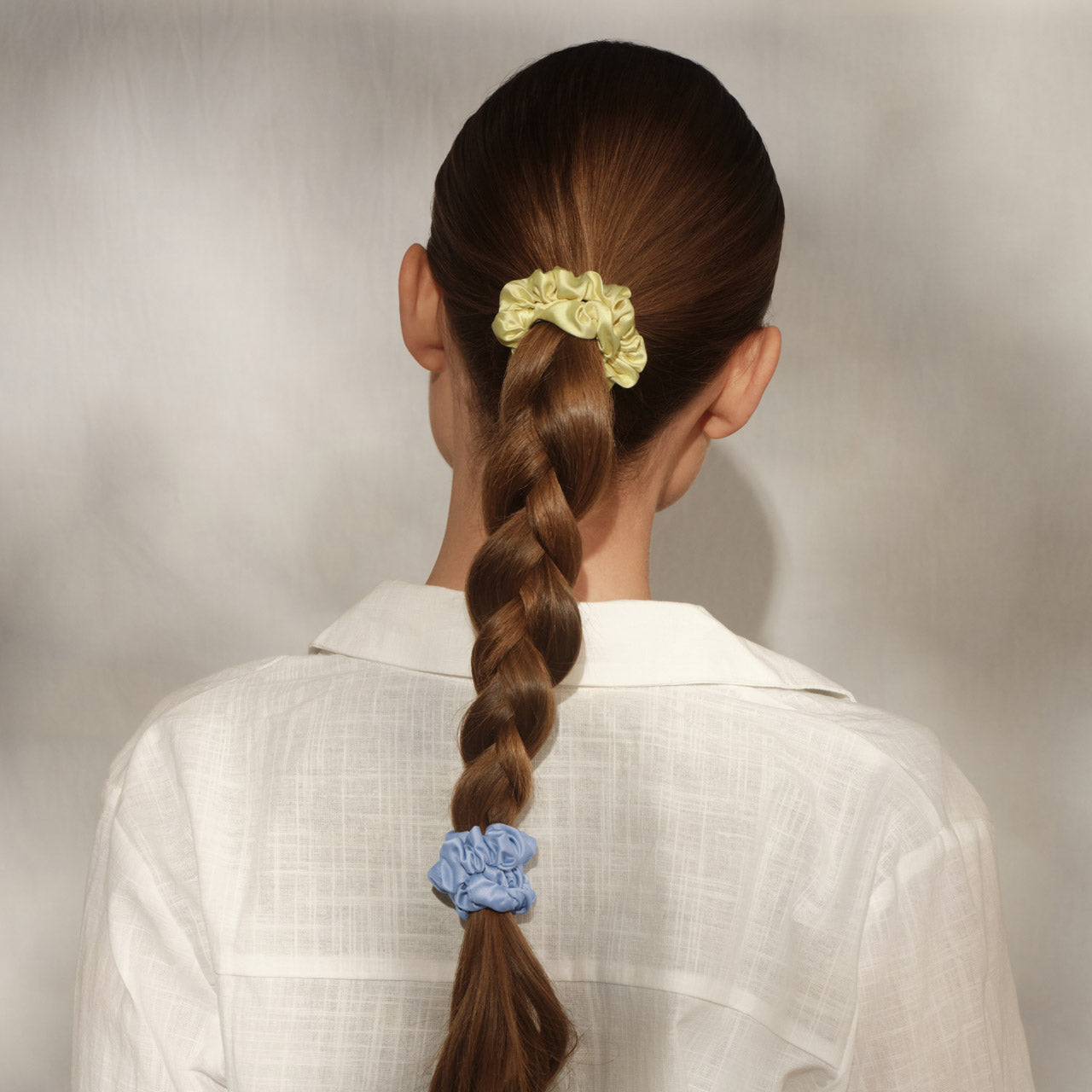 Back view of female model with long braided hair wearing KITSCH Coastal Cottage Ruched Satin Scrunchies — butter yellow scrunchie at top of braid, lavender blue at end — white linen shirt — Extra Gentle, snag-free hold for all hair types