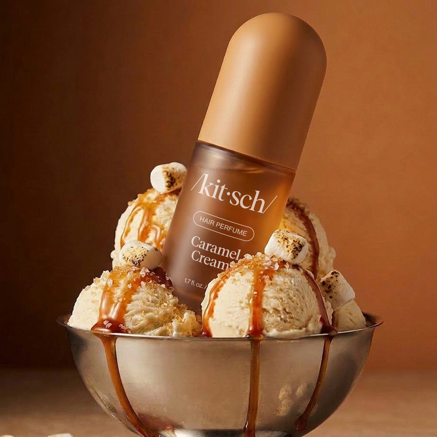 A Kit-sch Caramel Cream perfume bottle stands upright in a metal bowl filled with vanilla ice cream, caramel drizzle, and toasted marshmallows, against a warm brown studio background.